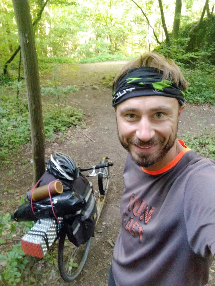 Muž s čelenkou a úsměvem na lesní cestě vedle naložené jízdní kolo s brašnami a přilbou
