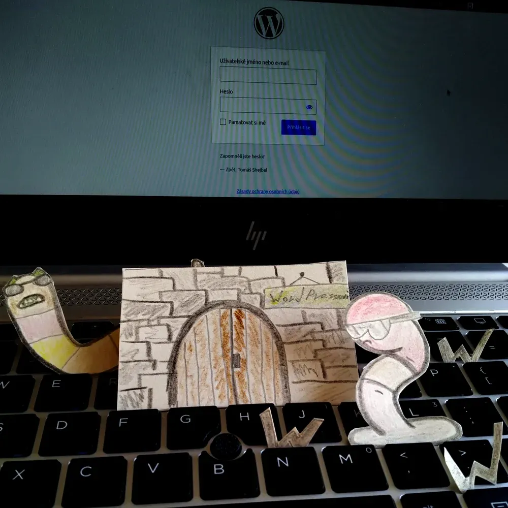 Vystřižené kreslené postavičky červů a papírová brána s nápisem "WordPressovi" stojí na klávesnici před přihlašovací obrazovkou WordPressu